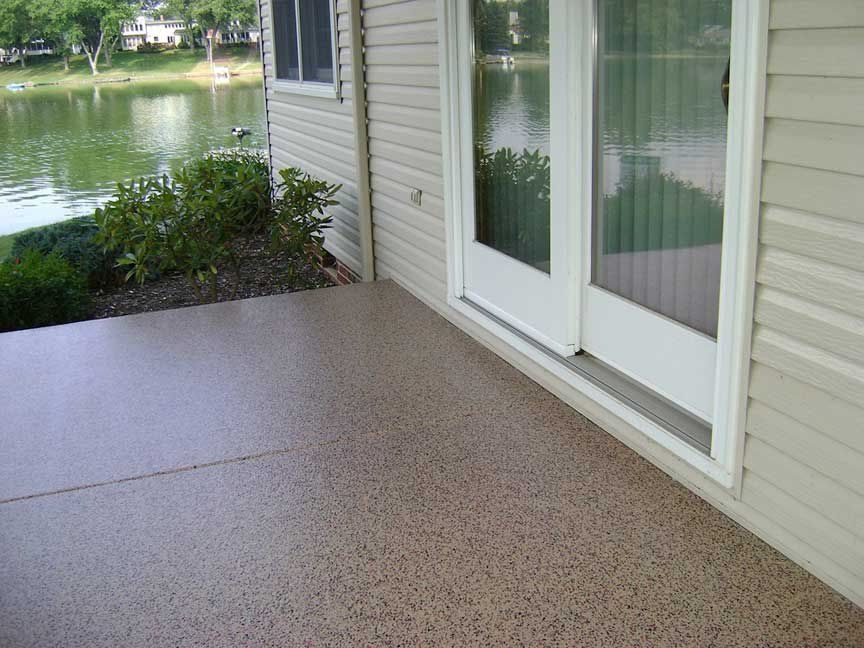 Patio with speckled floor, near a lake, next to a home with sliding glass door.