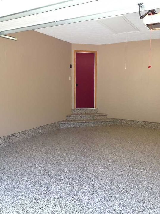 Empty garage with epoxy floor, tan walls, and a maroon door up a few steps.