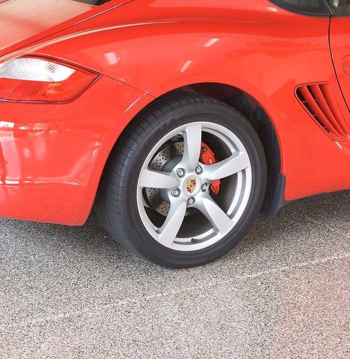 Red Porsche sports car rear wheel. Silver wheel, red brake caliper, gray tire, on a speckled floor.