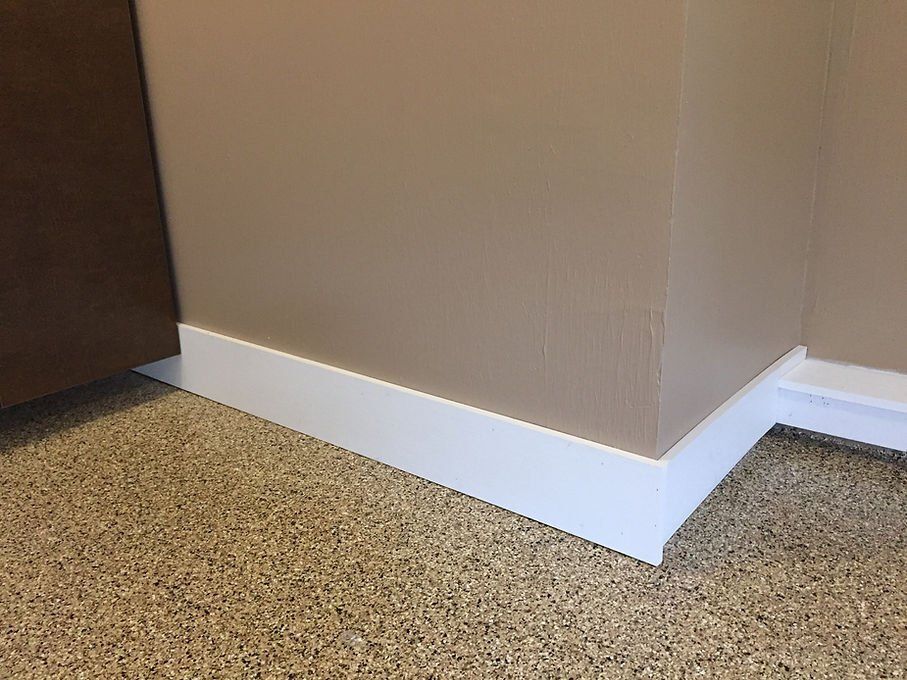 White baseboard molding at a corner, tan walls, speckled brown and tan flooring.