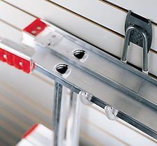 Ladder hanging on hooks attached to a white slotted wall, red handle detail.