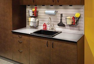 Garage sink with cabinetry, metal wall organizers, and cleaning supplies. Dark wood, gray countertop.