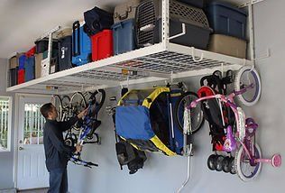 Man hanging bike in a garage with overhead storage holding boxes and pet carrier, bikes and stroller hanging below.