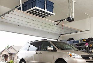 A car parked in a garage with overhead storage shelves holding blue bins. The garage door is partially open.