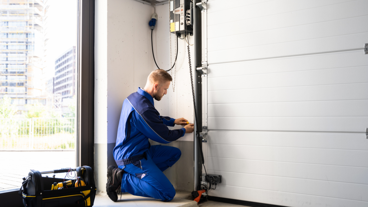 Person in blue coveralls fixing a white garage door, indoors. A toolbox sits on the floor.