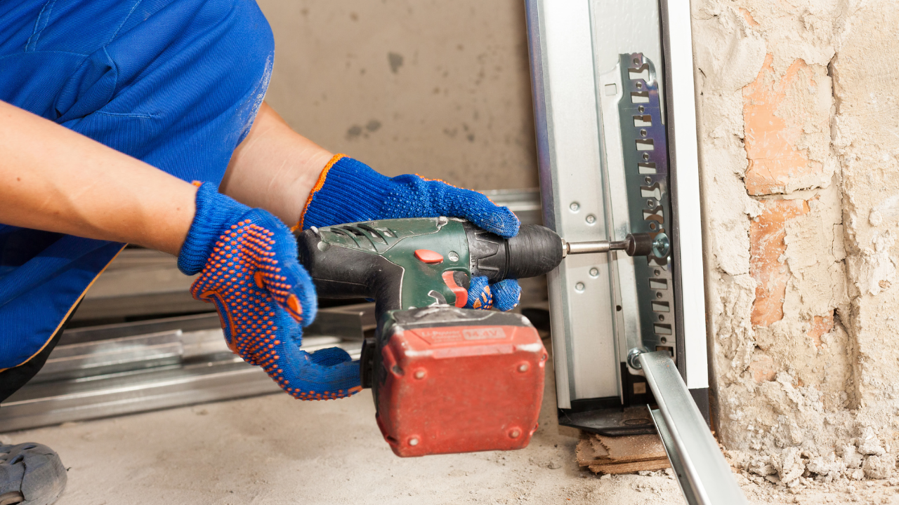 Person in blue work gloves using a drill on garage door track in a concrete setting.