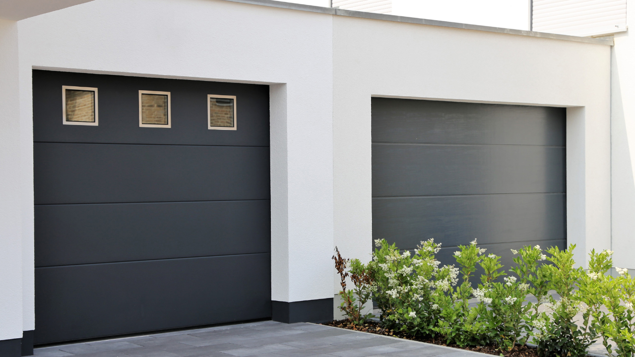 Two charcoal gray garage doors on a white building, small bushes in front.