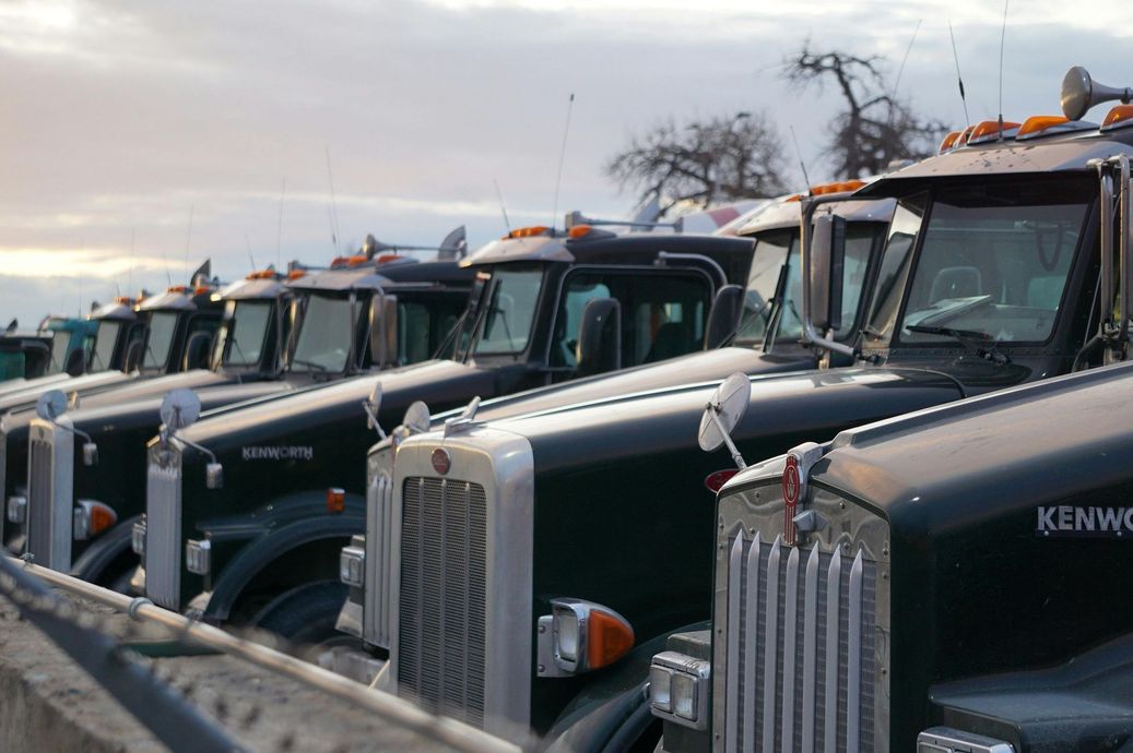 Row of parked Kenworth semi trucks in a lot, front grilles facing forward under a cloudy sky