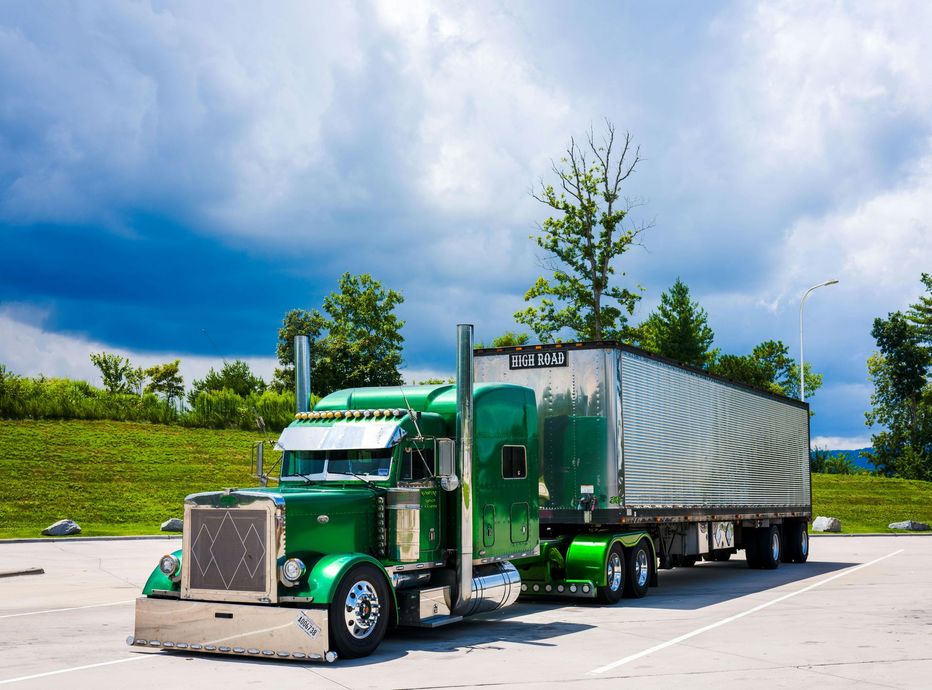 Green semi-truck with silver trailer parked in a lot under cloudy sky