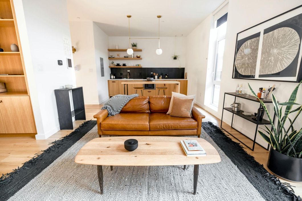 A modern living room with a brown leather sofa, wooden coffee table, large patterned rug, and open kitchen in the back.