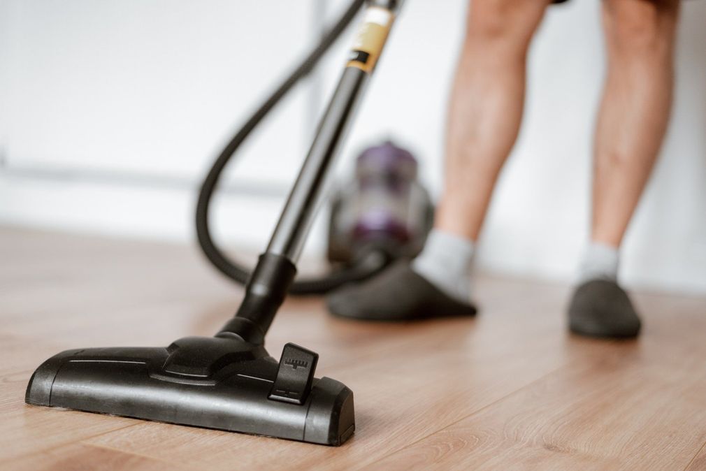 A person in slippers vacuums a light wood floor with a black canister vacuum.