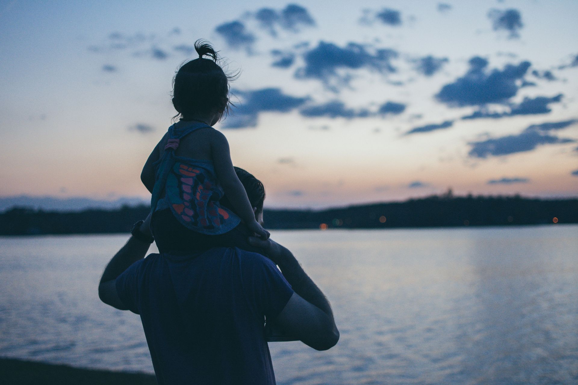 Person carrying a child on shoulders by a calm lake at dusk