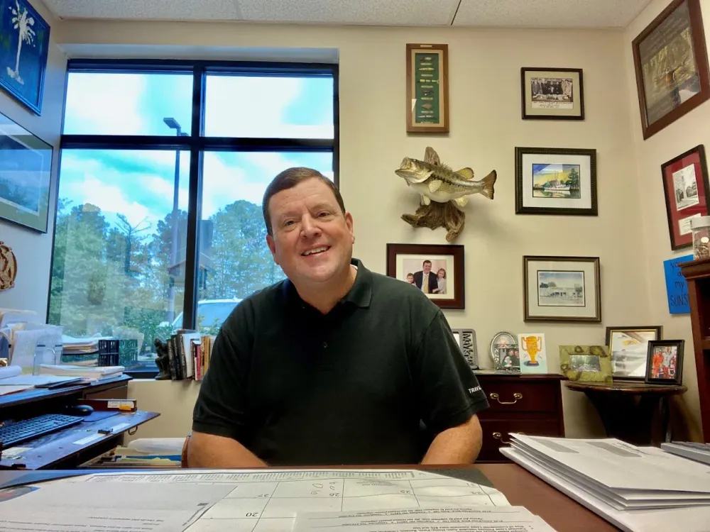 Smiling man seated in an office with framed art, a window, and papers on the desk