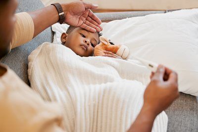 Person checking a child's forehead with hand, child wrapped in a blanket, holding a stuffed animal, indoors.