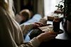 Person holding a tray with a glass of dark liquid. Another person is lying in bed in the blurred background.
