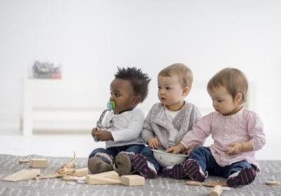 Three babies sitting on a rug with toys, one using a pacifier, the others looking at a bowl and blocks.