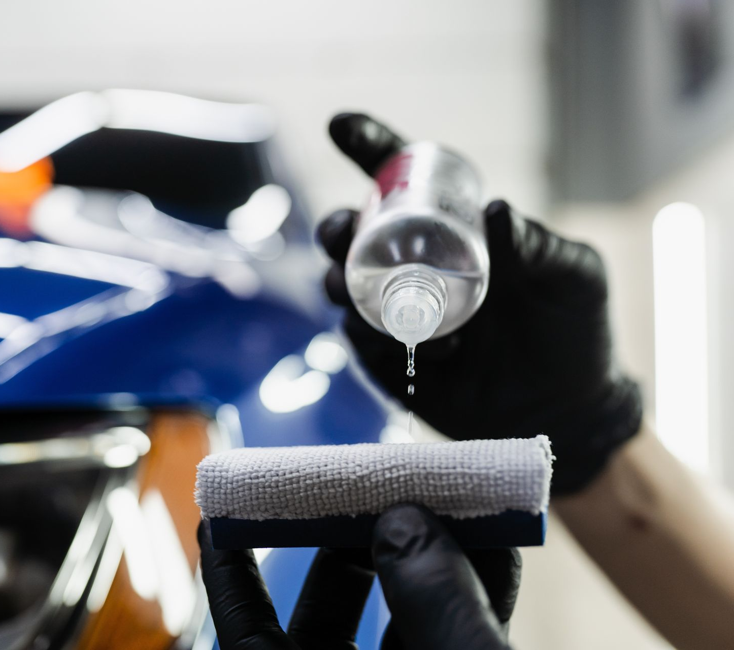 Person wearing black gloves applying liquid from a bottle to a detailing pad on a blue car.