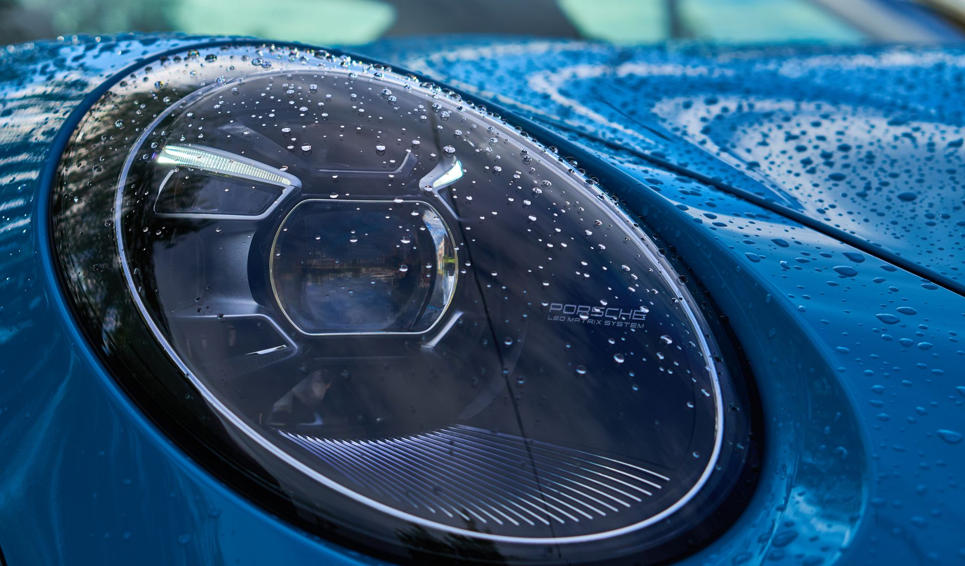 Blue Porsche headlight with water droplets on the lens, close-up.