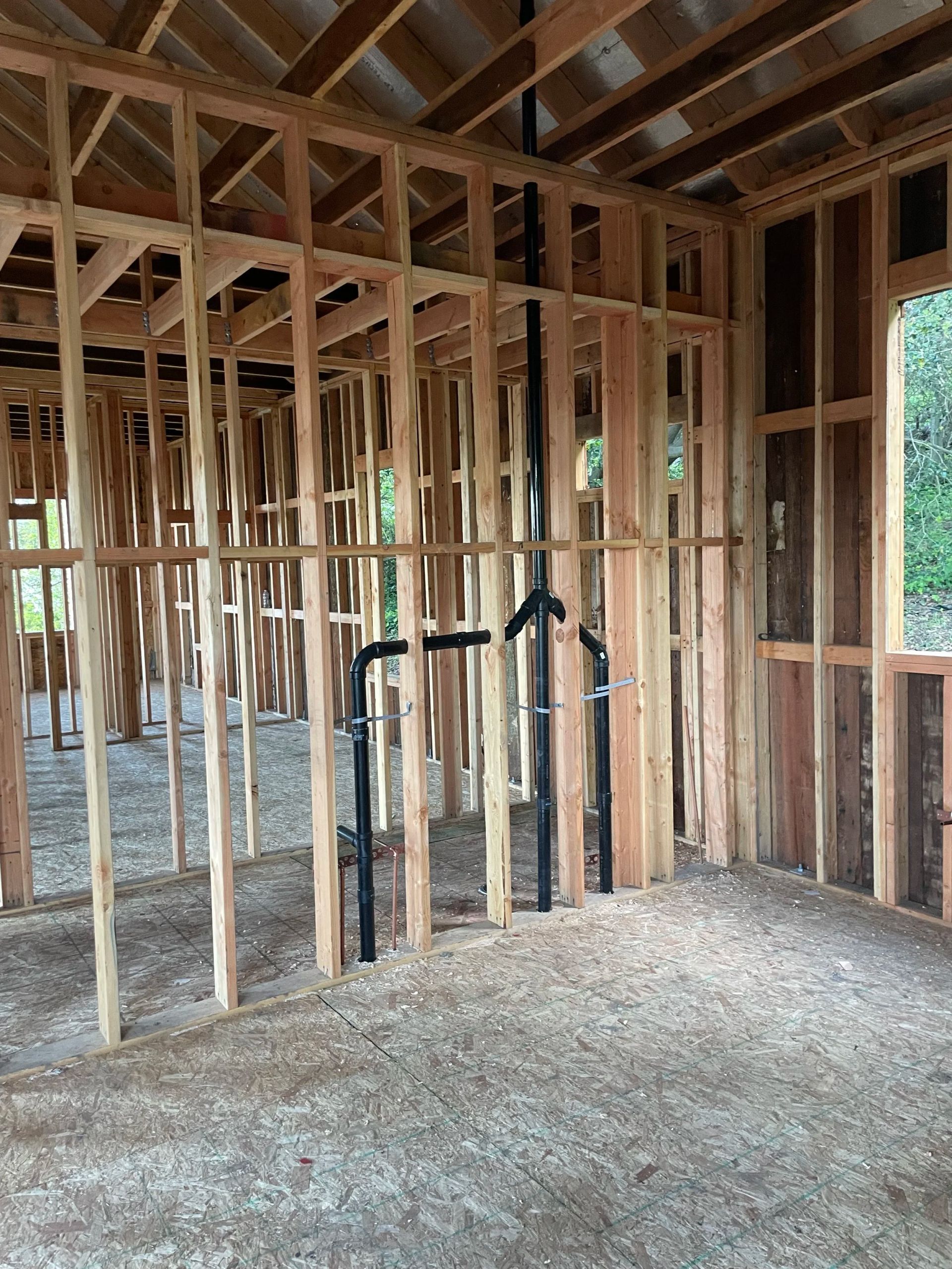 Unfinished house interior with exposed wooden framing, gravel floor, and plumbing pipes.