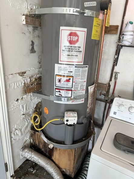 Gray water heater in a utility closet beside a washing machine and exposed pipes.