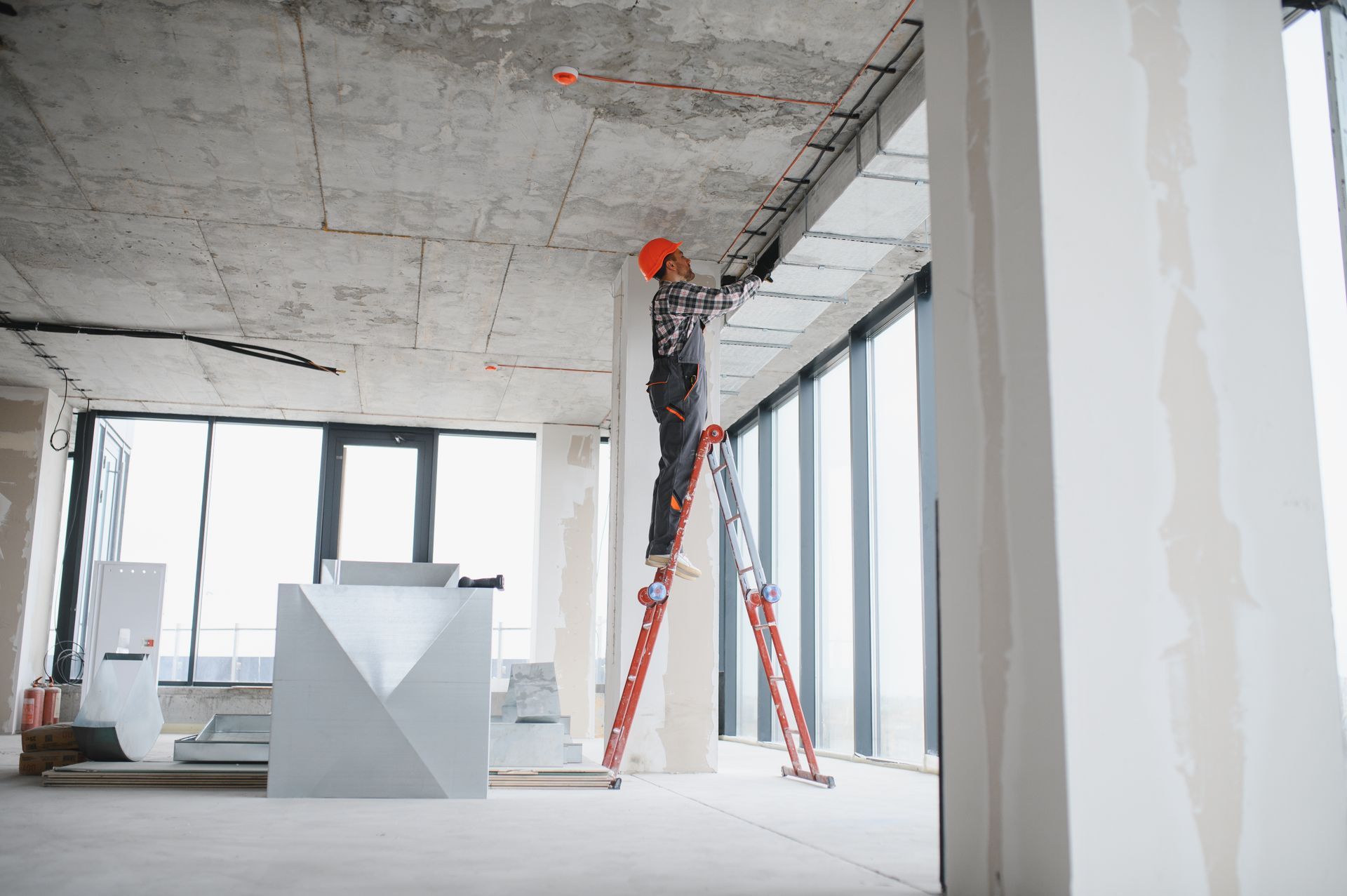Renovated apartment interior with concrete ceiling, large windows, ladders, and a worker near the wall