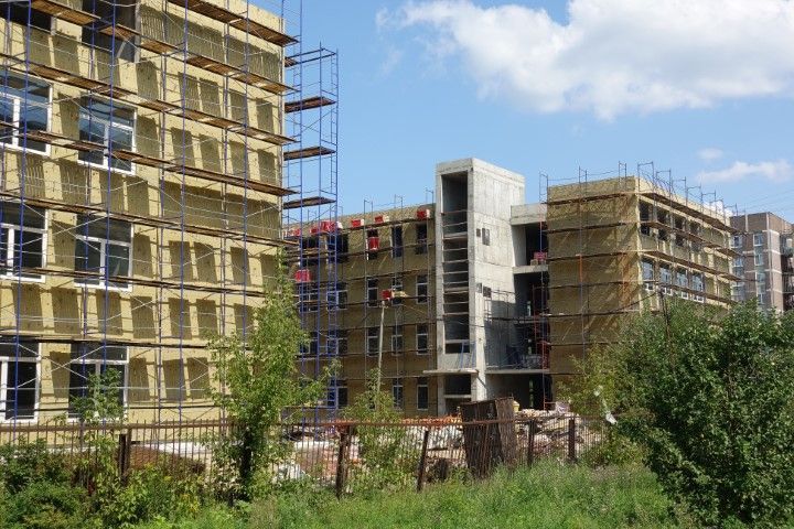Buildings under construction with scaffolding, covered in yellow insulation, against a cloudy sky.