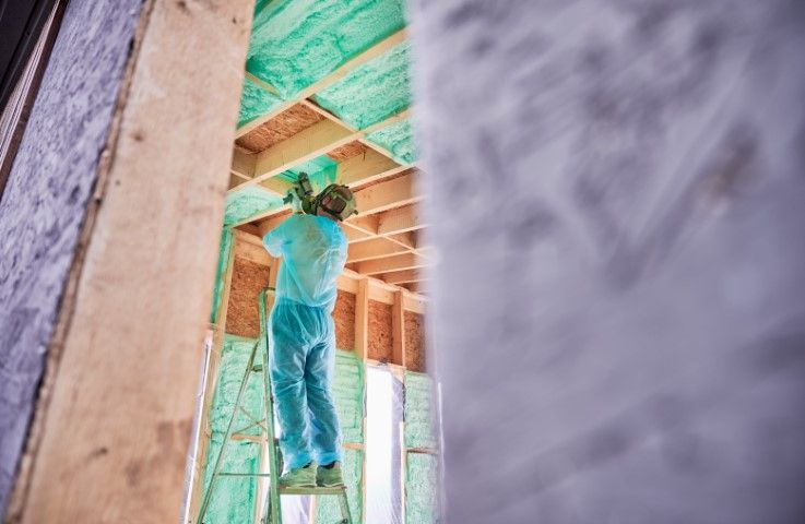 Worker on a ladder spraying insulation into the framing of a building; interior view.