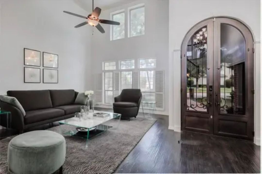 Bright living room with dark sofa, glass coffee table, armchair, ceiling fan, and arched wooden doors.