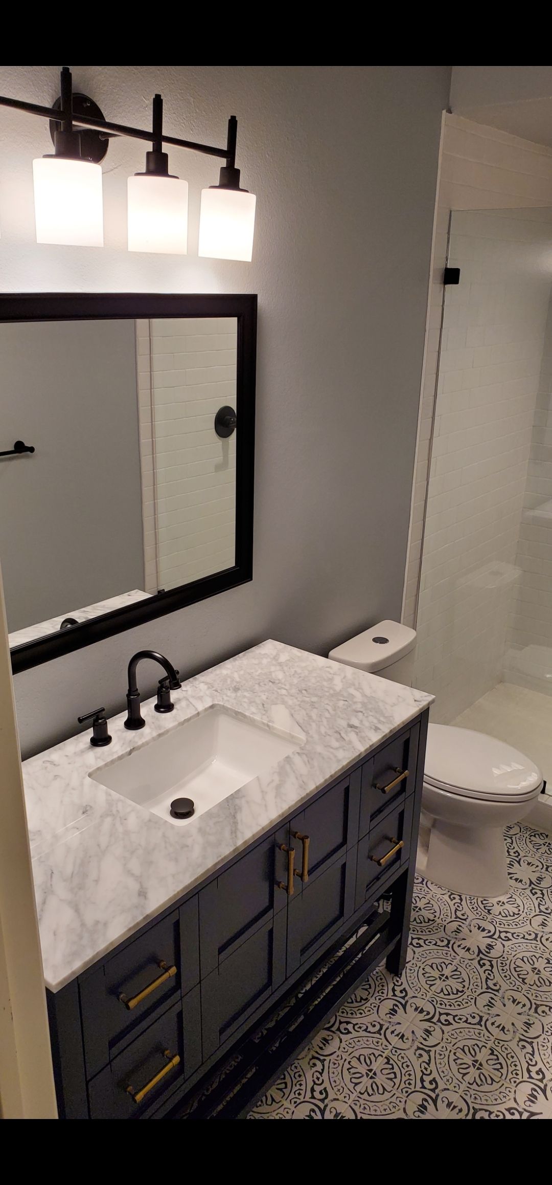Modern bathroom with dark vanity, white marble sink, mirror, wall lights, toilet, and tiled floor