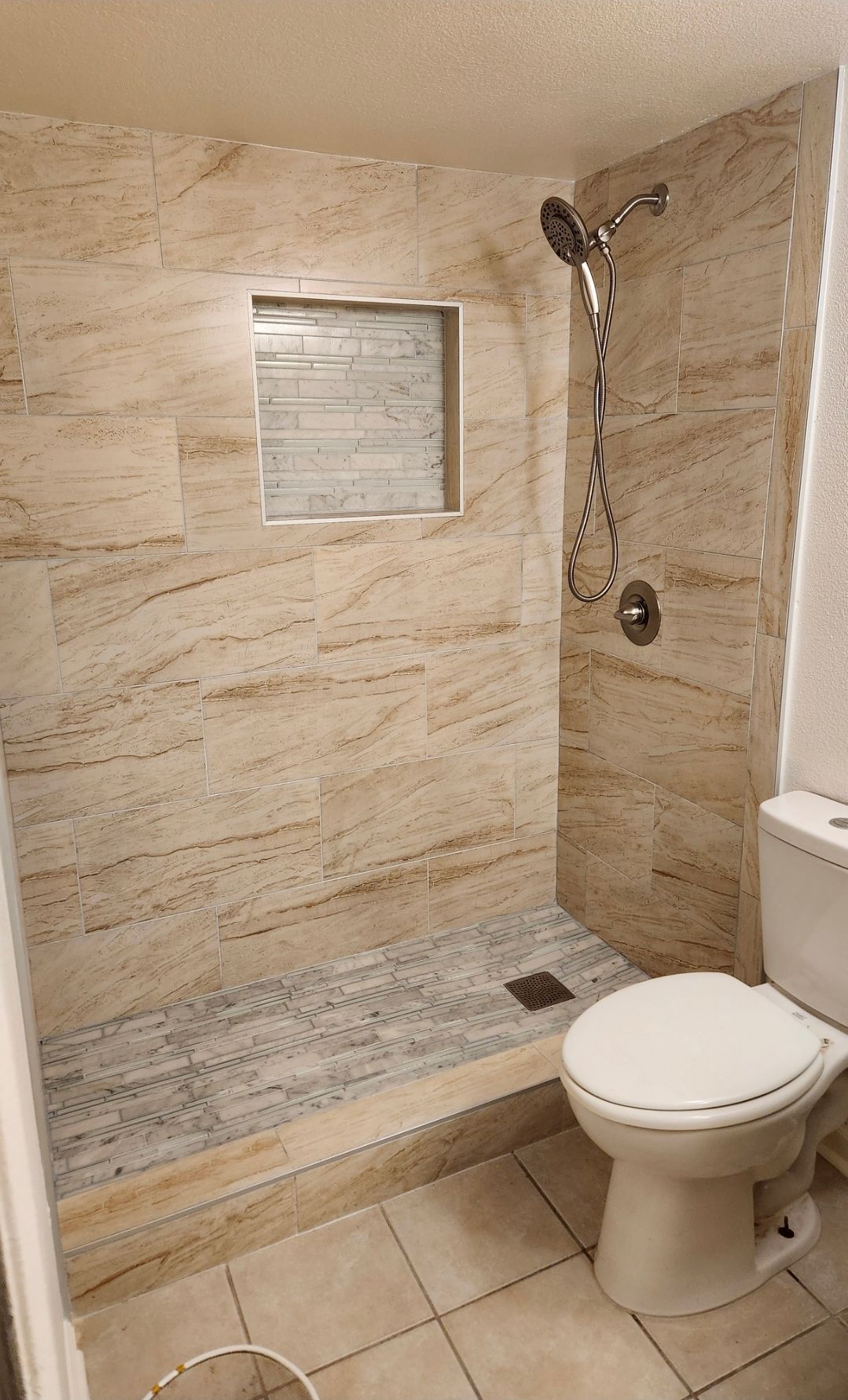 Small beige tiled bathroom with walk-in shower, wall niche, handheld showerhead, and toilet in foreground