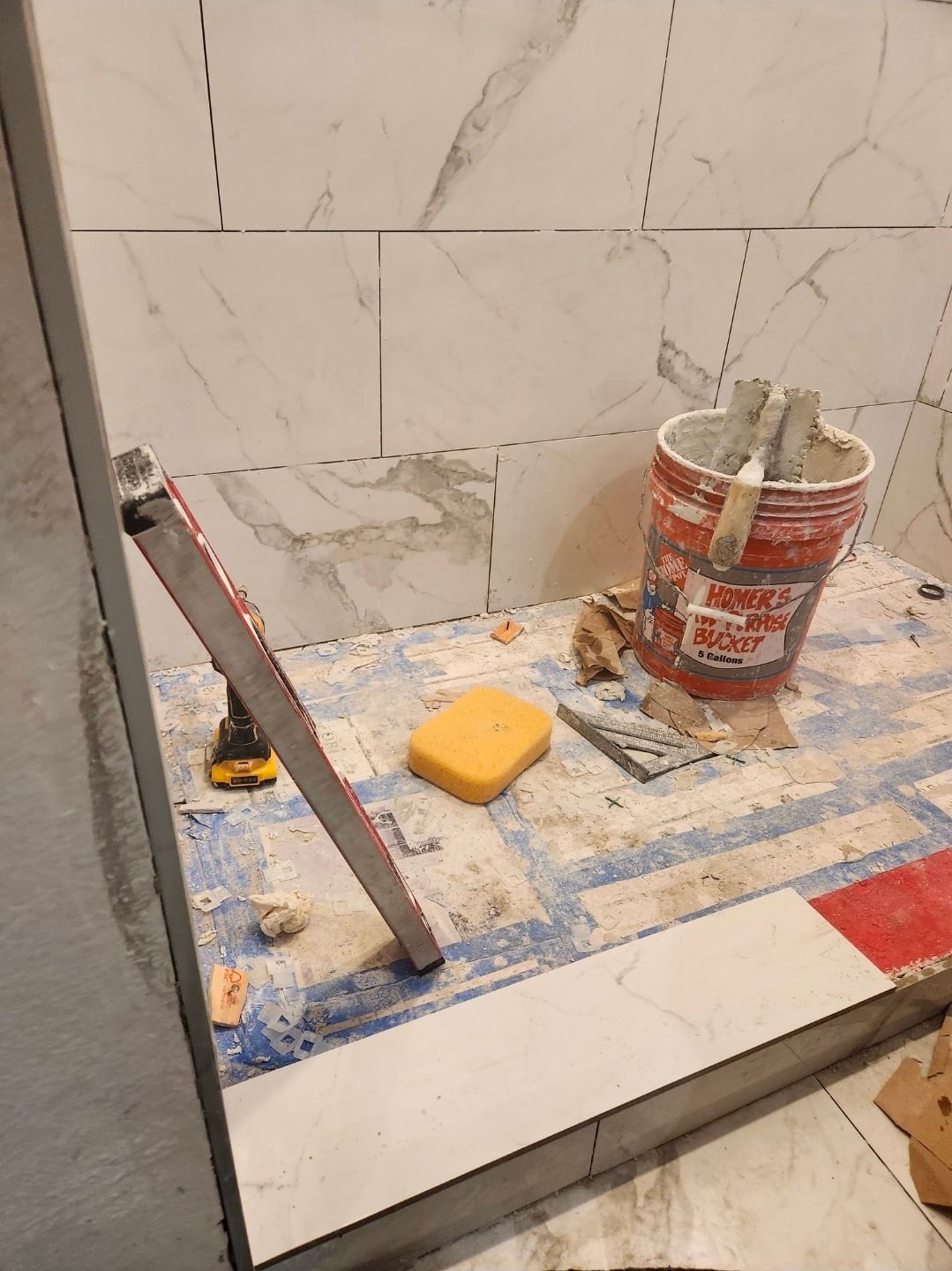 Tiled shower floor under construction with red leveling tool, bucket, and sponge on a blue protective surface