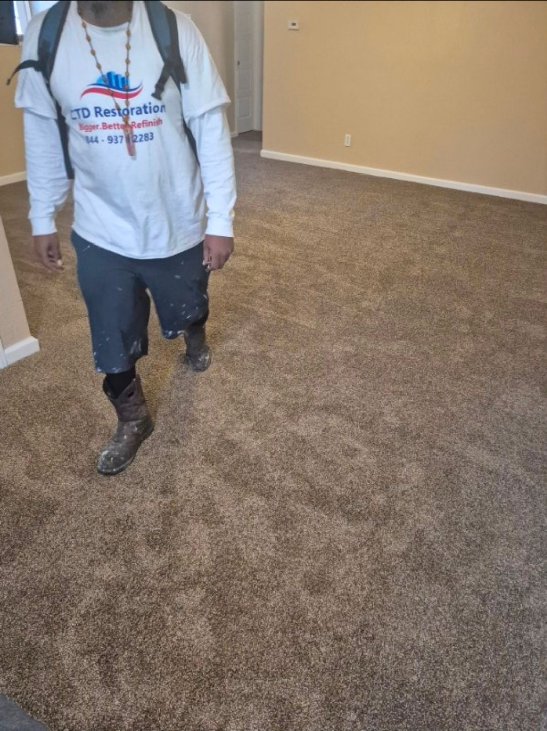 Person in a white shirt and dark overalls walking across a carpeted room.