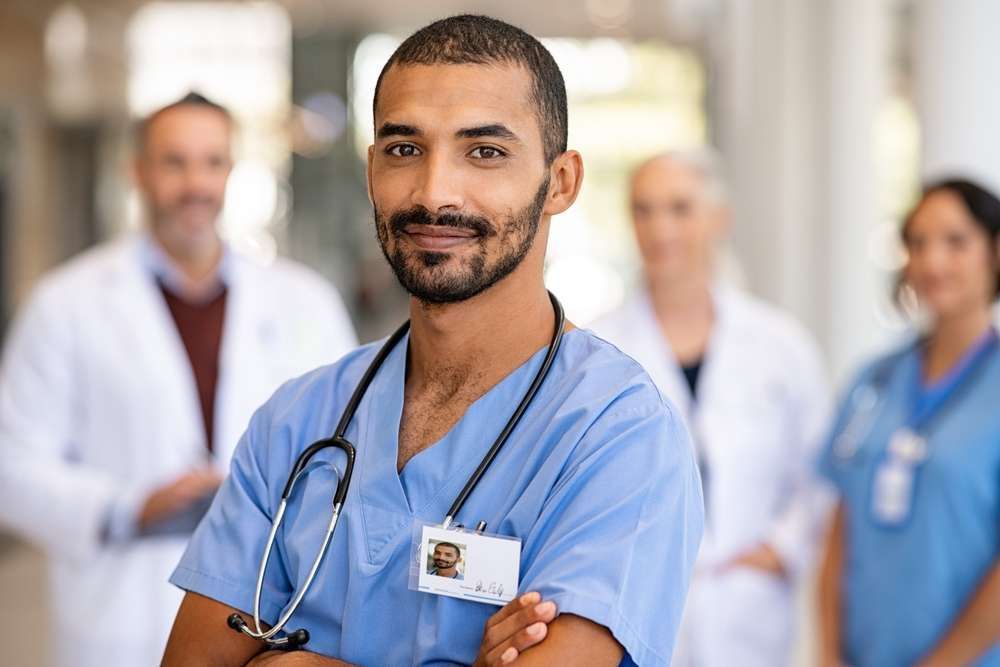 Healthcare worker in blue scrubs with stethoscope, smiling, arms crossed, in a hospital hallway.
