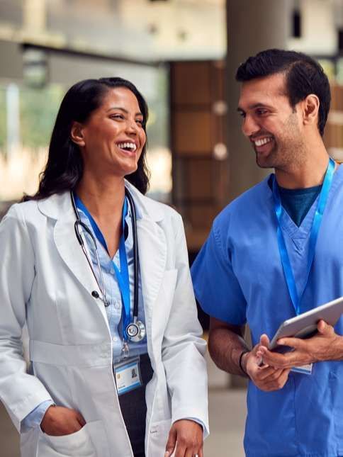 Two medical professionals, smiling, walking in a hospital hallway. One wears a white coat, the other blue scrubs, both have lanyards.