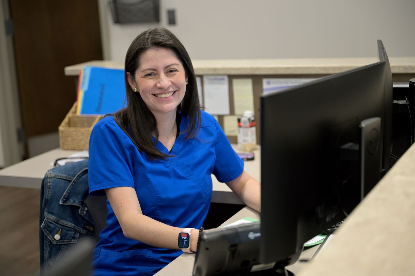 Woman in blue scrubs smiles at a desk, looking at a computer monitor.