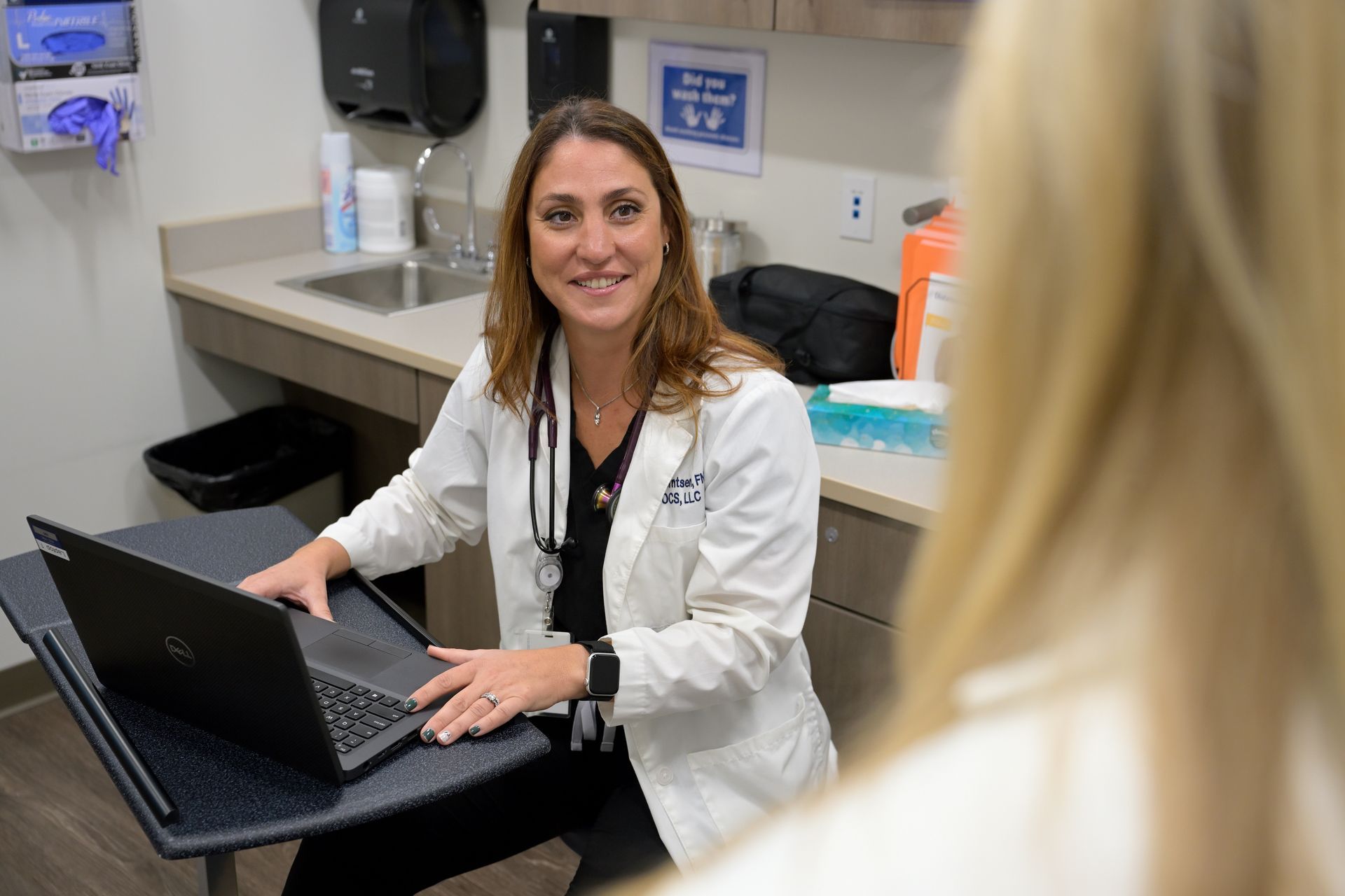 Doctor in lab coat smiling at a patient while using a laptop in an exam room.