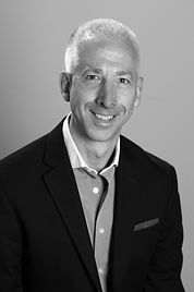Smiling man in blazer, button-down shirt, neutral background.