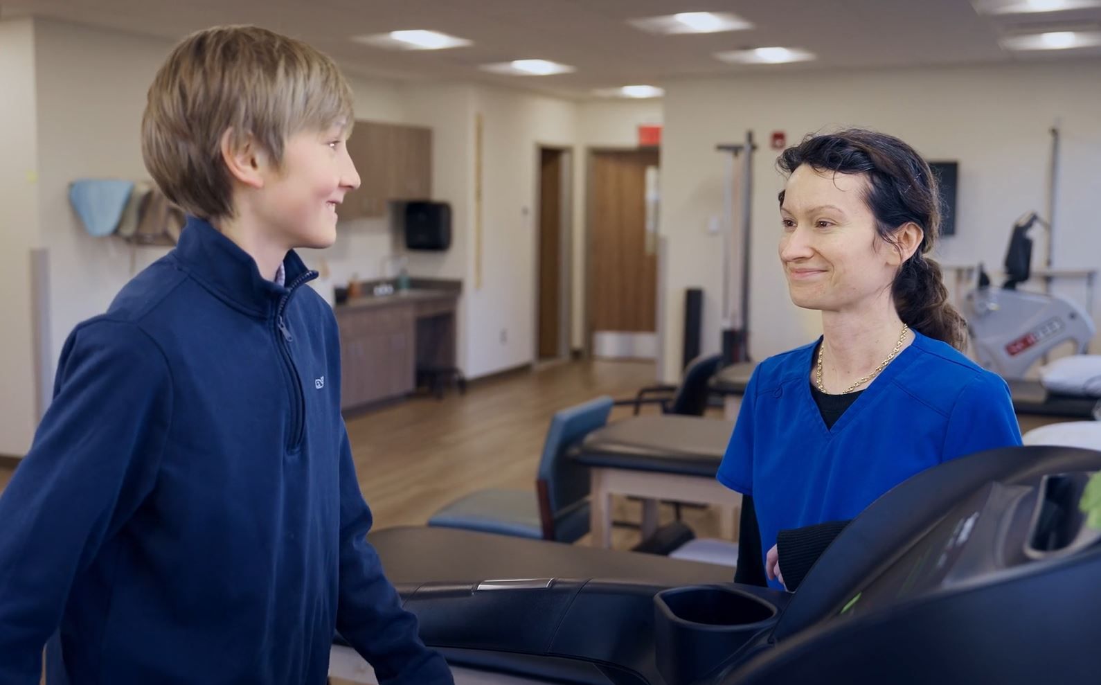 A person in blue jacket smiles at a person in blue scrubs near a treadmill in a physical therapy room.