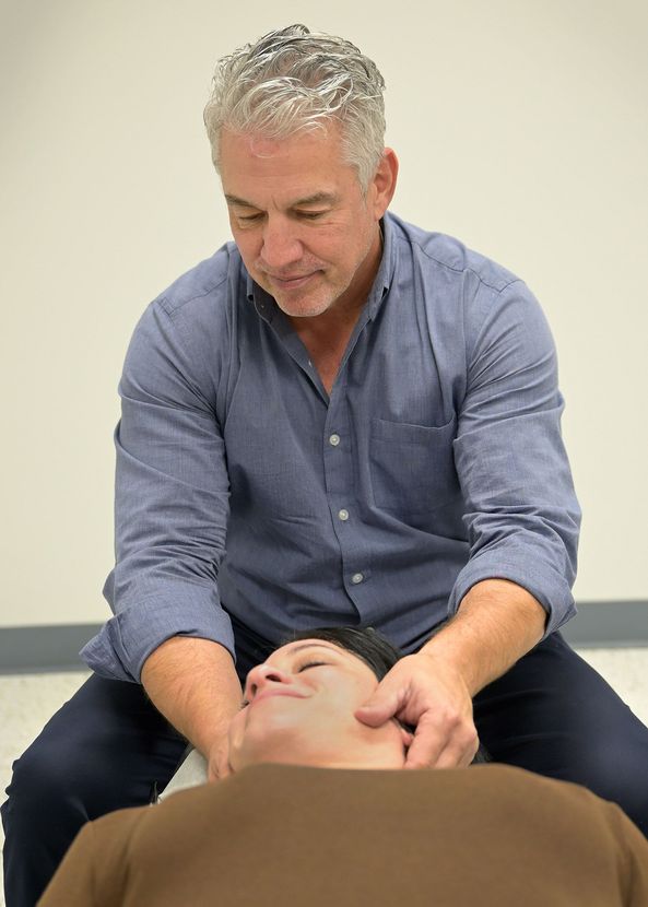 Man performing neck manipulation on a person, indoors. He's wearing a blue shirt. The other person is lying down.