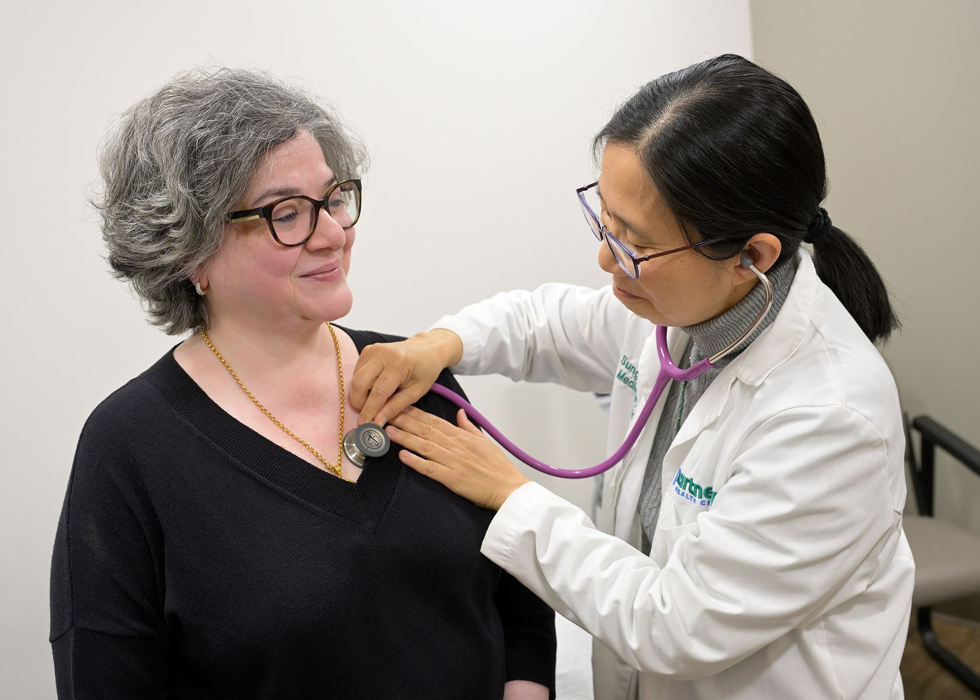 Doctor using a stethoscope on a patient's chest. Indoors, medical setting. Patient in black sweater, smiling. Doctor in white coat.