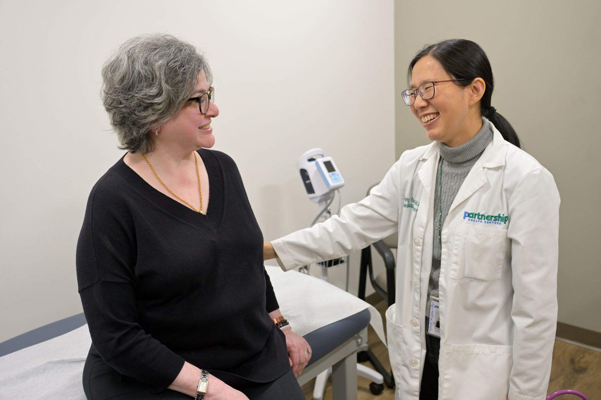 A doctor in a white coat smiles at a seated patient in a medical examination room.