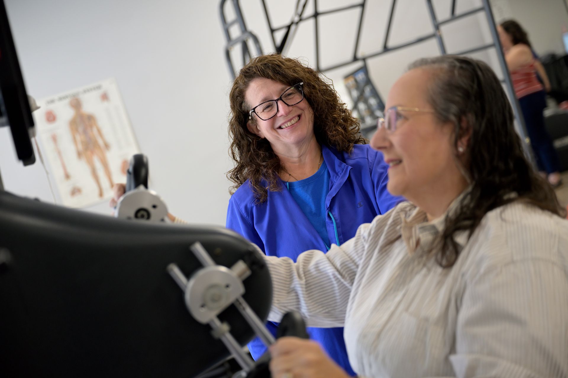Woman assisting another woman using exercise equipment, smiling in a rehabilitation setting.