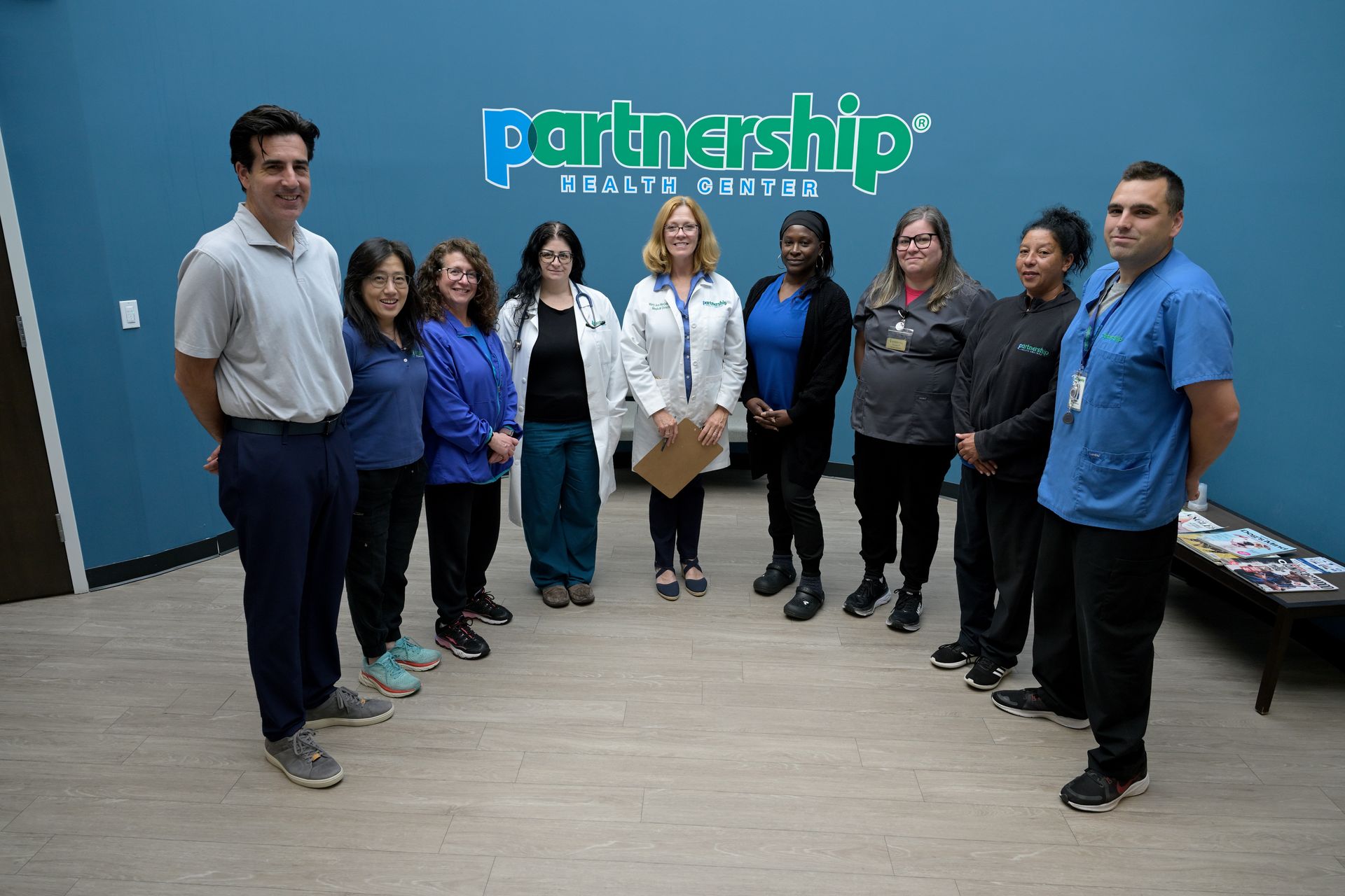 A group of nine healthcare professionals stand side-by-side in front of a blue wall with the Partnership Health Center logo.