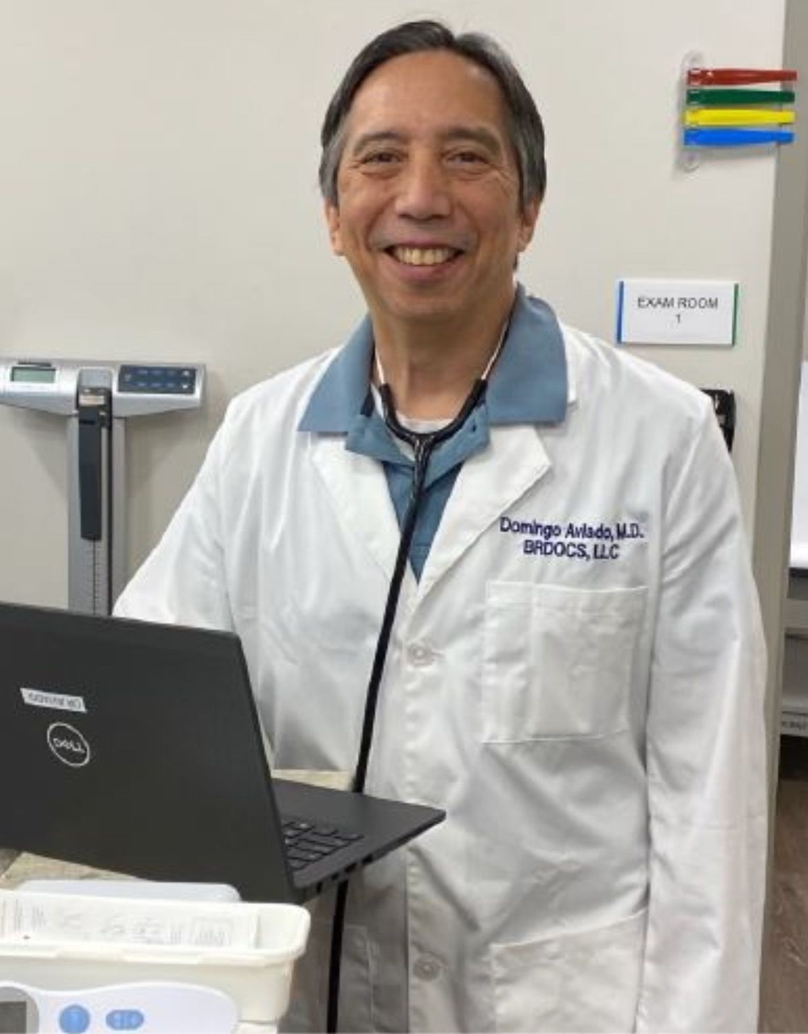 Doctor with stethoscope and laptop, wearing white coat, smiling in an exam room.
