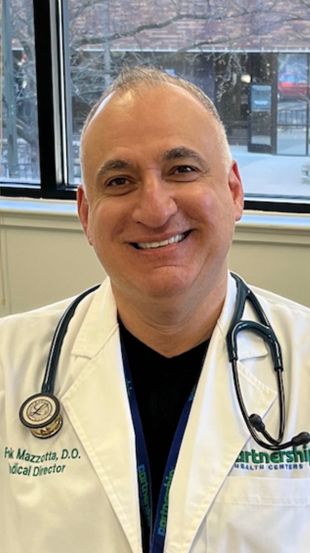 Man wearing a white coat with a stethoscope around his neck, smiling. The background is a window.