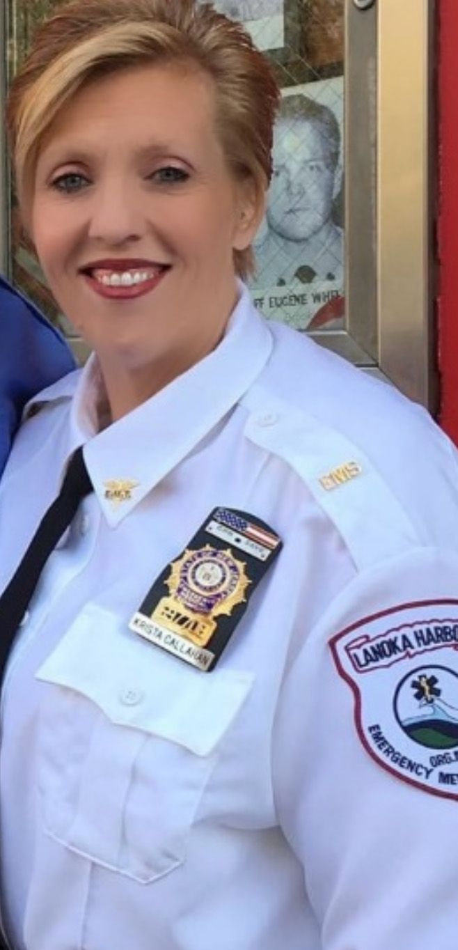 Woman in white uniform smiling, wearing a badge, near an emergency services emblem.