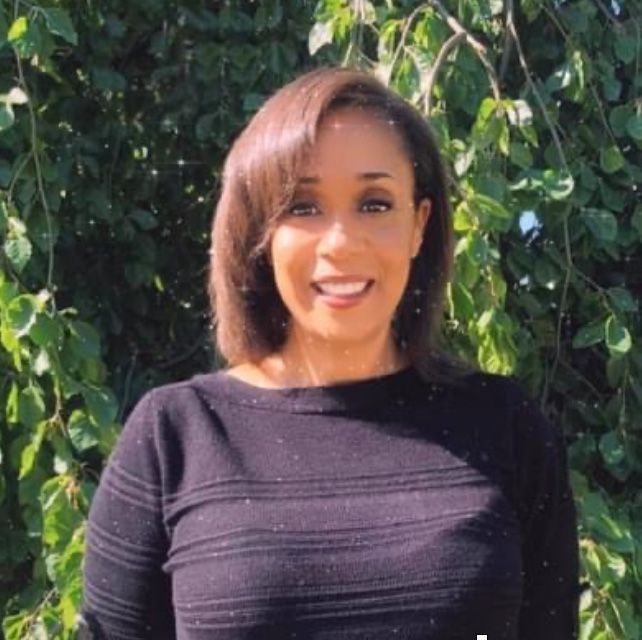 Woman smiling, wearing a black sweater, set against a background of green foliage.