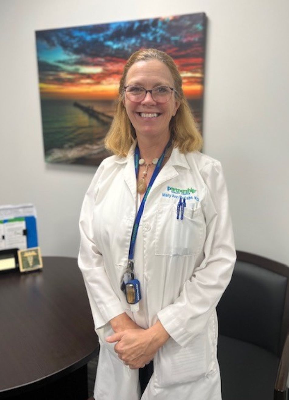 Woman in white lab coat smiles; in office setting with art, desk.