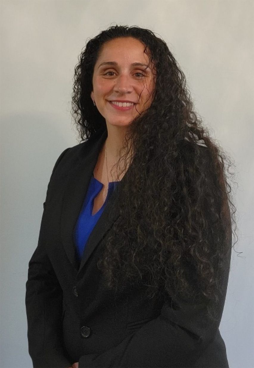 Woman with long, curly hair smiles, wearing a blue top and black blazer against a light gray wall.