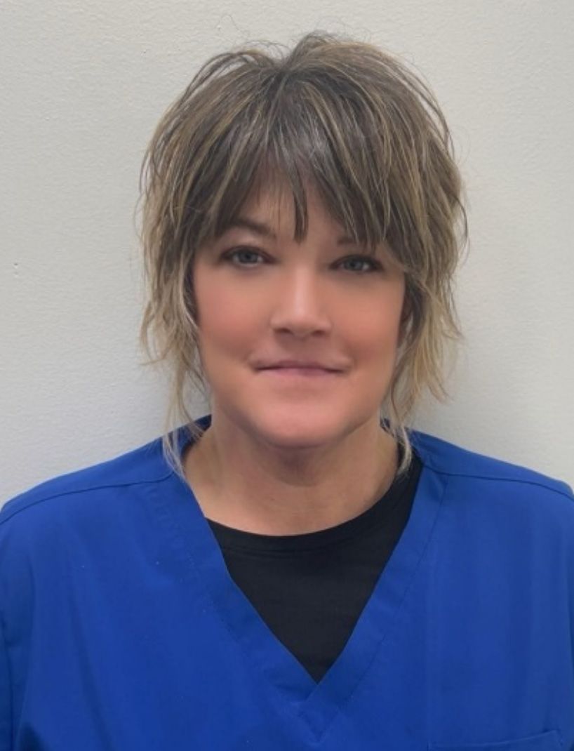 Woman with short, layered hair, wearing a blue scrub top over a black shirt, smiling slightly.